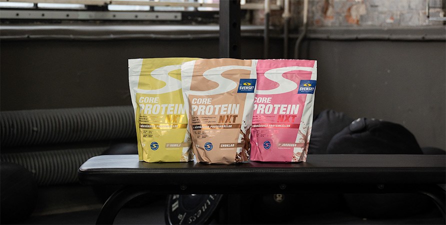 Tre påsar Core Protein NXT från Svenskt Kosttillskott i smakerna vanilj, choklad och jordgubb står på en träningsbänk i ett gym.