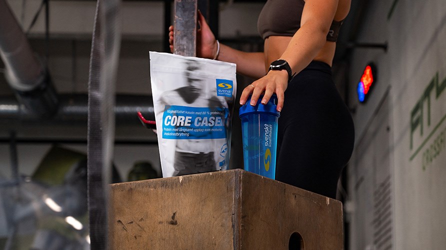 En kvinna i träningskläder håller handen på en shaker som står bredvid en förpackning Core Casein på en trälåda i ett gym. En kvinna i träningskläder håller handen på en shaker som står bredvid en förpackning Core Casein på en trälåda i ett gym.