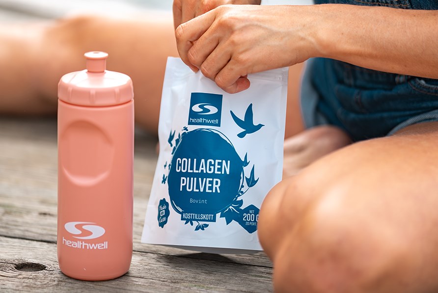 Healthwell Collagen Pulver Bovint 2.0 visas i nrbild p en brygga, tillsammans med en vattenflaska frn Healthwell. En kvinnas ben r synliga men r inte i fokus. 