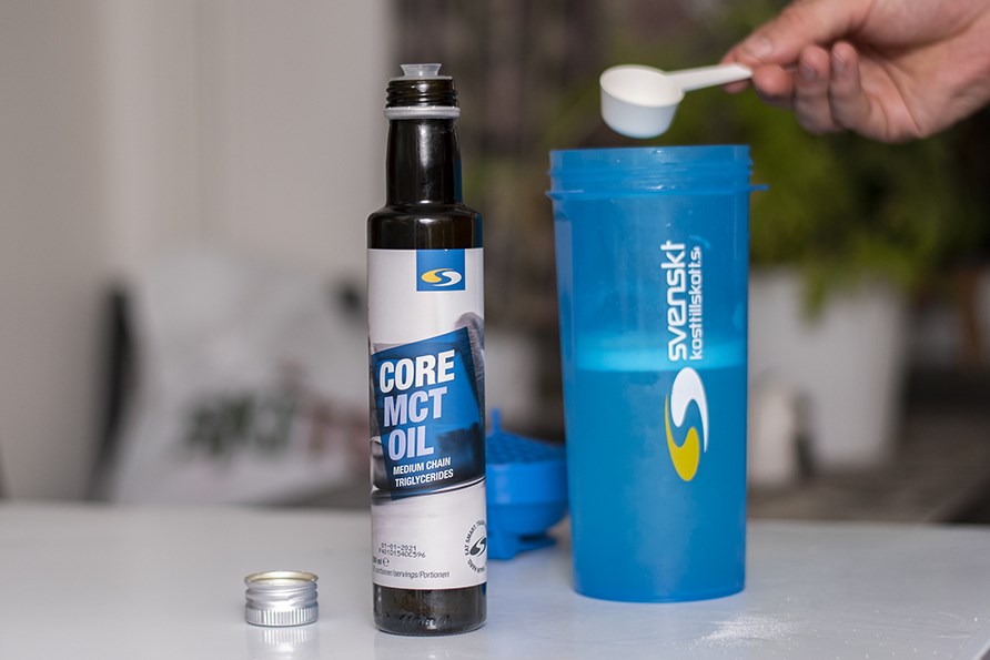 Bild på Core MCT-olja i flaska, en shaker i bakgrunden och en hand som häller 1 msk MCT-olja i shakern.
