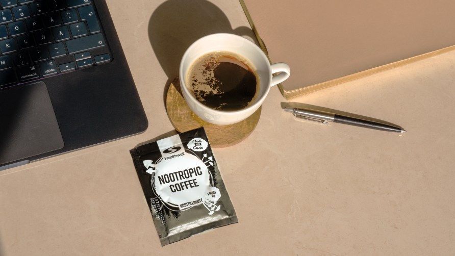 En pse Healthwell Nootropic Coffee ligger p ett skrivbord med dator och en kopp kaffe.  