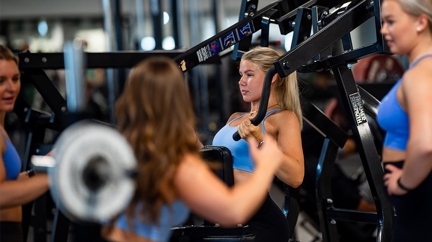 Fyra unga kvinnor pratar med varandra samtidigt som de anvnder trningsmaskinerna p ett gym.